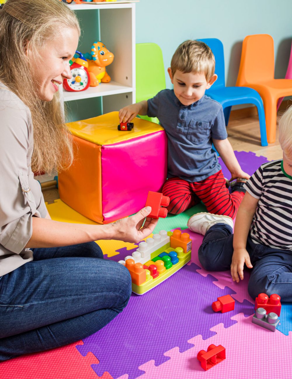 Two little boys are playing with a kindergarten teacher. The woman is sitting with boys on the floor in the playroom